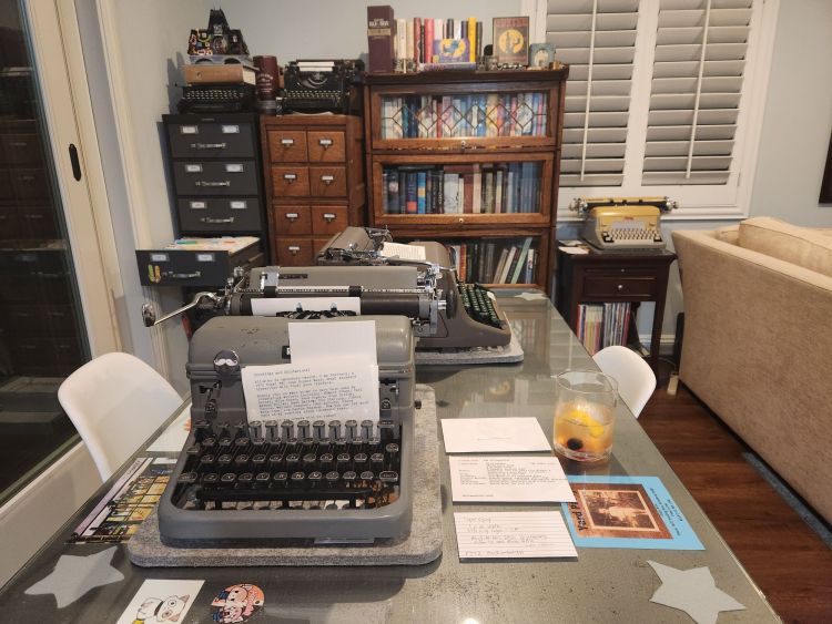 Desk set up with a gray Royal KMG standard typewriter and a brown Royal HH. Next to the KMG is a cocktail glass with a yellowish whisky sour in it. In the background are two card index cabinets and a barrister bookshelf full of books. In the background we see three other typewriters.
