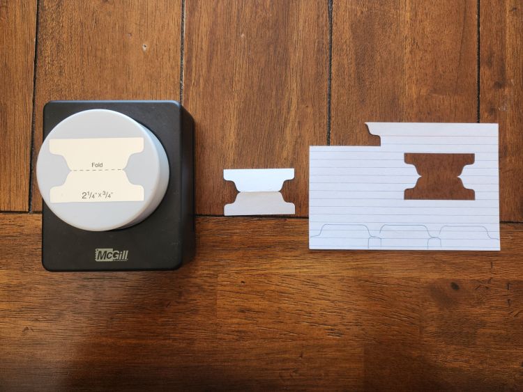 A square black plastic punch with a white circular button sits on a wooden table next to an index card with a tab shaped hole punched out of it.