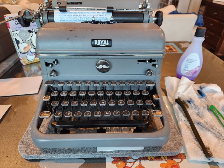 A gray Royal KMG typewriter on a felt mat sits on a table next to some index cards on one side and a purple bottle of nail polish remover (acetone), a brass bristle brush, and a toothbrush sitting on a blue-ink-stained paper towel on the other. A careful observer might see a black pin with a white moustache stuck to the front of the typewriter underneath the Royal badge.