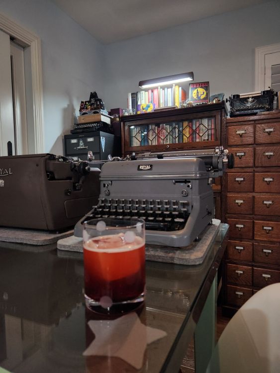 An old fashioned glass with a large square ice cube and an eerie , blood red cocktail chills in front of a large gray Royal KMG typewriter. In the background a work lamp illuminates a group of books on a book case.