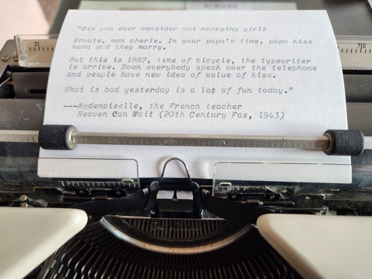 Typed index card in a typewriter carriage with a quote typed in italic face that reads: Did you ever consider not marrying girl? Ecoute, mon cherie. In your papa's time, papa kiss mama and they marry. But this is 1886, time of bicycle, the typewriter is arrive. Soon everybody speak over the telephone and people have new idea of value of kiss. What is bad yesterday is a lot of fun today.—Mademoiselle, the French Teacher / Heaven Can Wait (20th Century Fox, 1943)