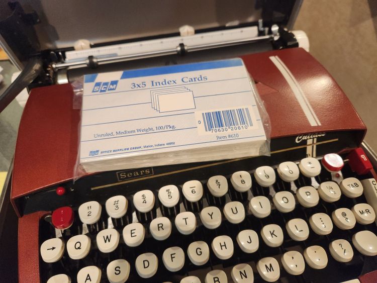 A pack of 100 3x5" index cards with the corporate logo SCM sits on top of a burgundy Sears Cutlass typewriter.