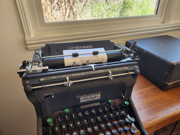 Black Underwood SS standard typewriter sitting on a wooden desktop in front of a window with greenery outside. There is a typed index card in the carriage.