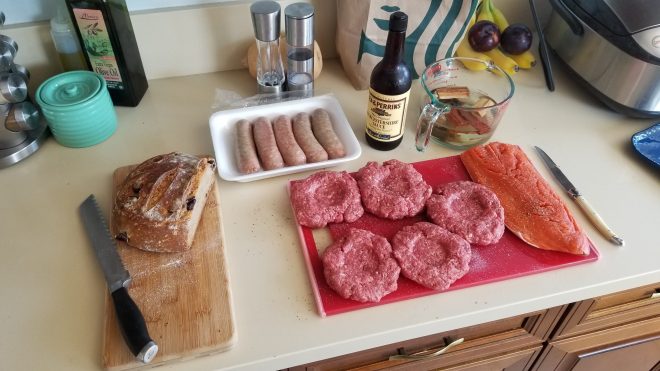 Counter with meat prepped to cook including 5 hamburgers, Italian sausages, and a large salmon steak
