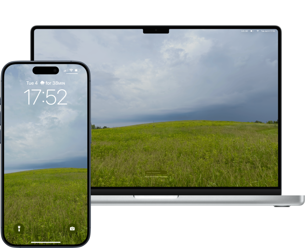 A MacBook and an iPhone display the same scenic wallpaper of a grass hill under a cloudy sky.