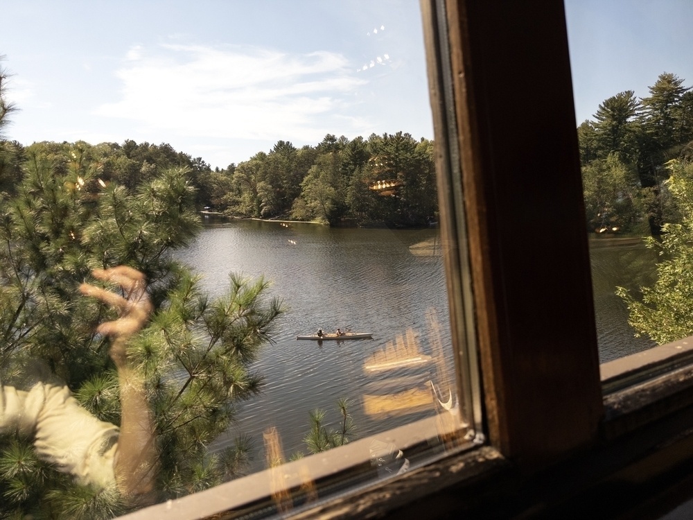 A view through a window of a serene lake surrounded by dense, green trees. The image captures two people in a canoe paddling across the calm water. The reflection of the person taking the photo, as well as the interior lights from inside, are visible in the glass, adding an overlay effect to the scene. The clear blue sky and lush greenery create a peaceful, natural setting.