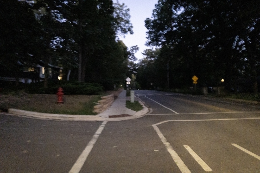 A dimly lit street scene at dusk, with a blurry and slightly out-of-focus presentation. The soft lighting creates an eerie and quiet atmosphere, with darkened trees and faintly visible houses lining the road.