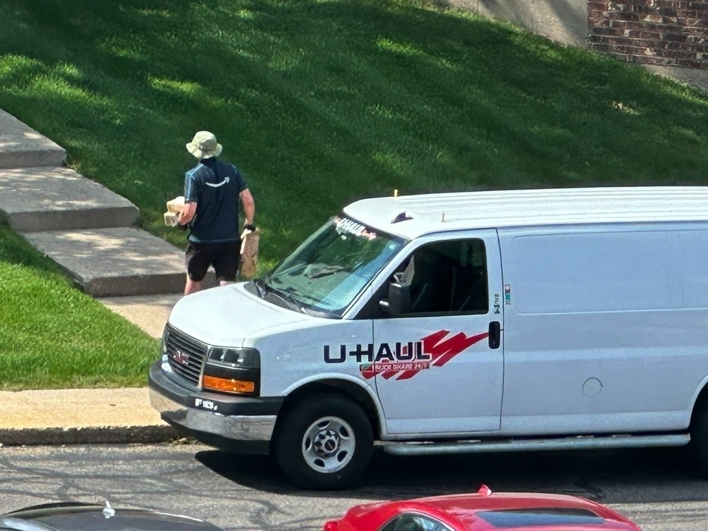 A man wearing an Amazon vest carries packages away from the U-Haul van in which he got them and up a path through grass