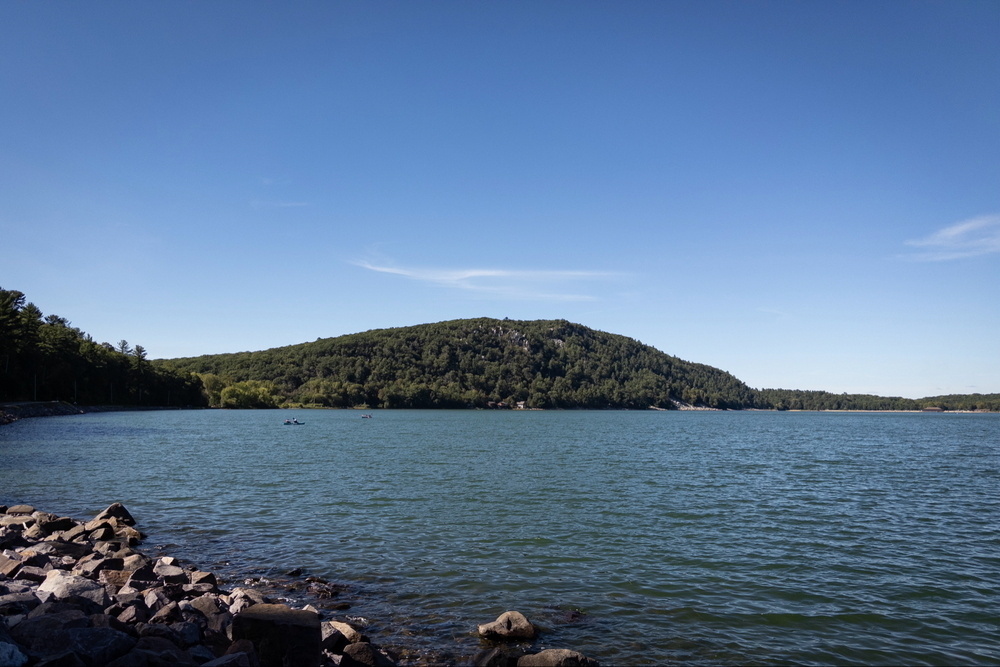 A serene lake with calm blue waters, bordered by a rocky shoreline on the left and surrounded by lush green hills under a clear blue sky.