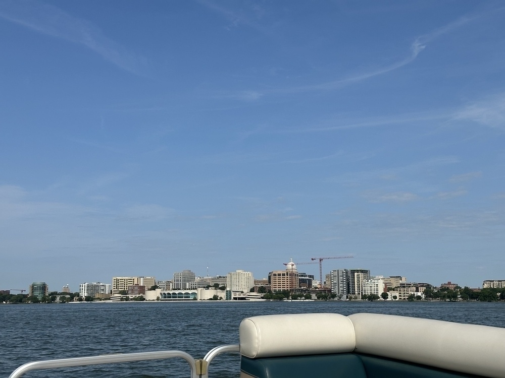 A city skyline viewed from a boat on a lake, featuring a blue sky with thin clouds and various buildings.