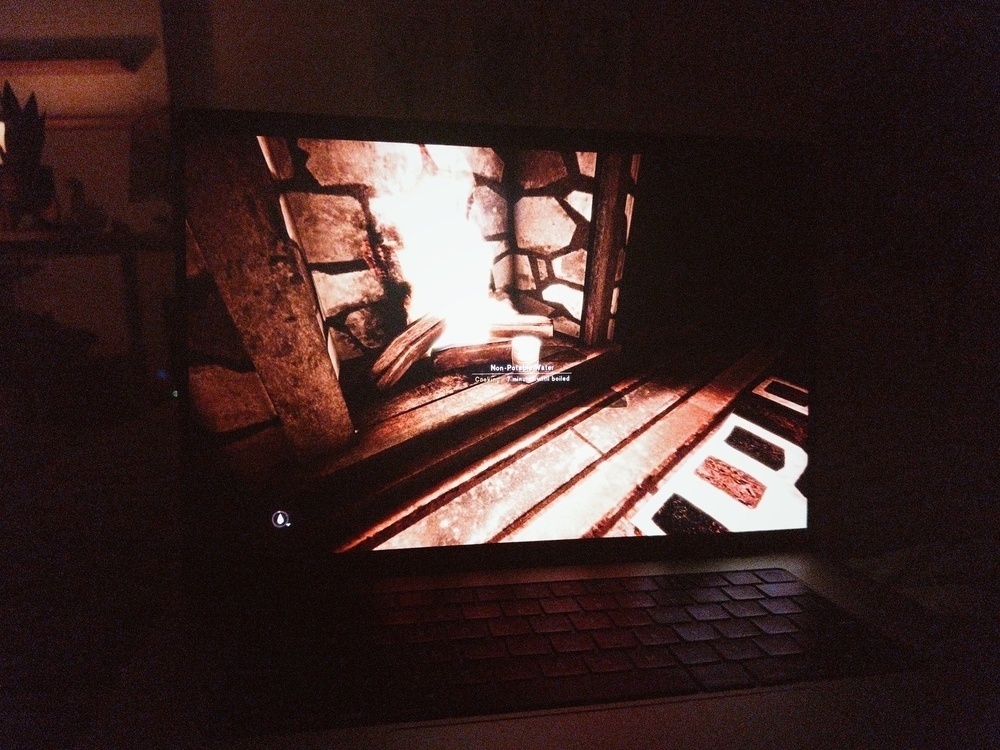 The photo shows a dimly lit room with a MacBook displaying the video game "The Long Dark." The screen depicts an in-game fireplace with warm, glowing embers and stone detailing. The rest of the room is dark, emphasizing the light from the laptop screen. The environment in the game is cozy, matching with the dimly lit room surrounding the laptop.