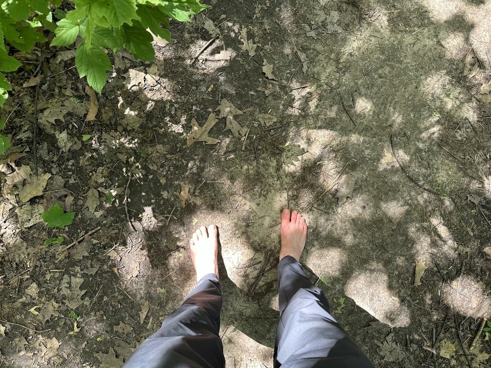 Me standing barefoot on a dirt trail. Sun is shining on my feet and there is foliage to the side.