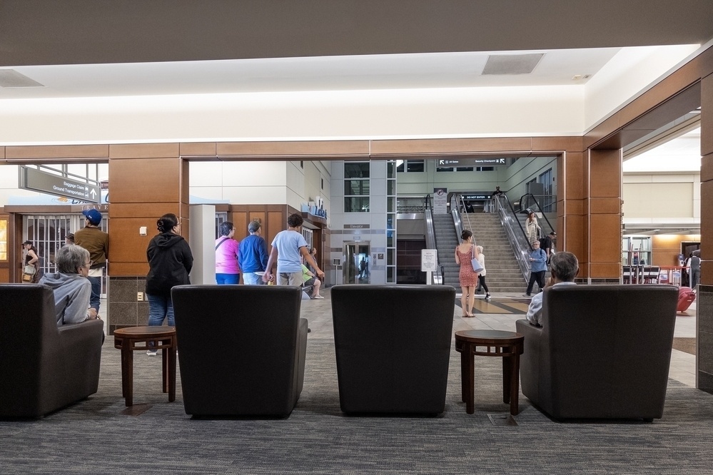 People are walking and sitting in an airport terminal, with some seated in lounge chairs in the foreground, facing an escalator and elevator in the background. Signs above direct travelers to baggage claim and ground transportation.