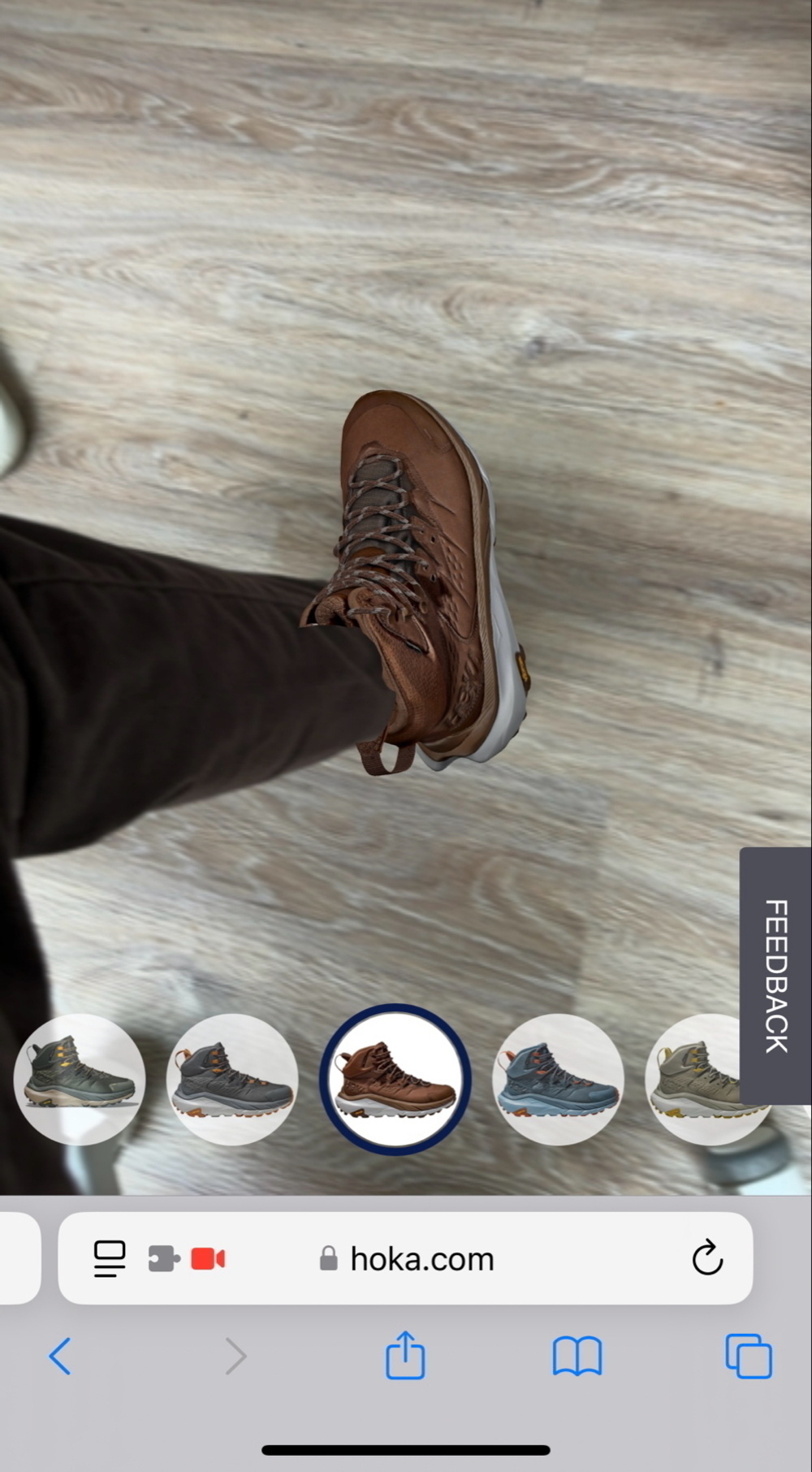 A person is trying on a brown hiking shoe using augmented reality (AR) on their mobile device — the shoe is shown virtually overlaid on their foot. The interface suggests the user is browsing shoes on the Hoka website and experimenting with different color options through an AR feature.