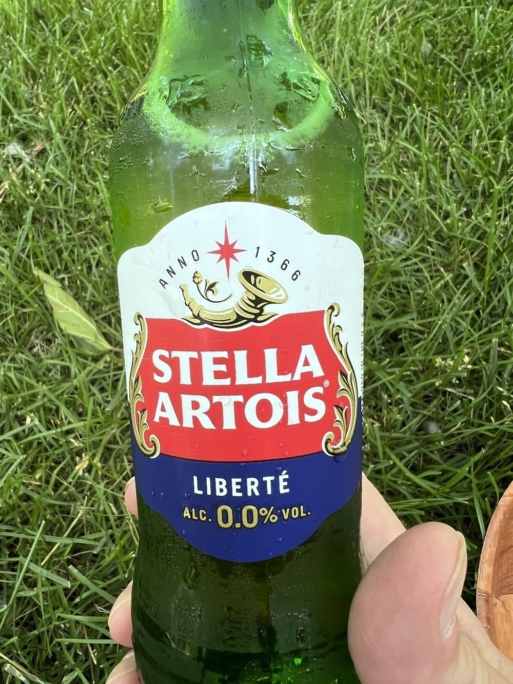A hand is holding a bottle of Stella Artois Liberté, which is a non-alcoholic beer with 0.0% alcohol content, in front of a grassy background.