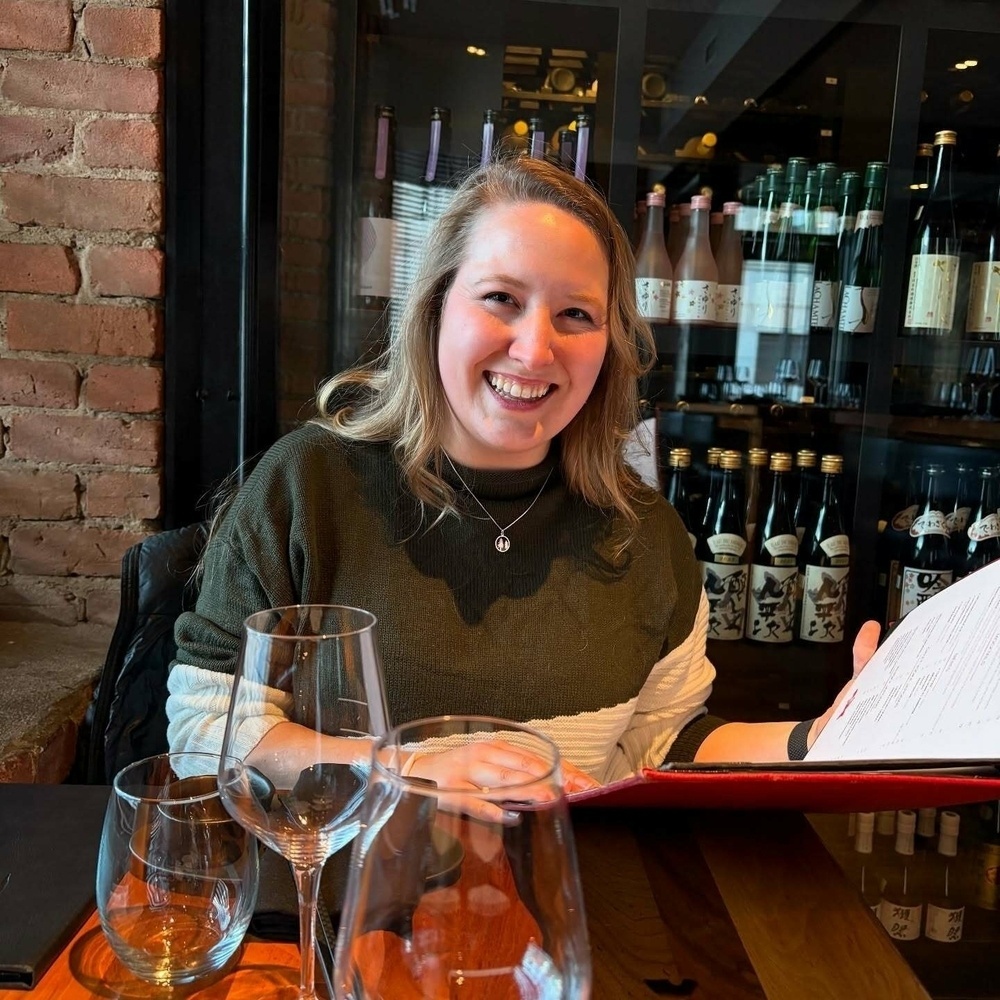 A woman smiling while holding a menu sits at a table with empty glasses in a restaurant. Behind her, shelves display numerous bottles with various labels.