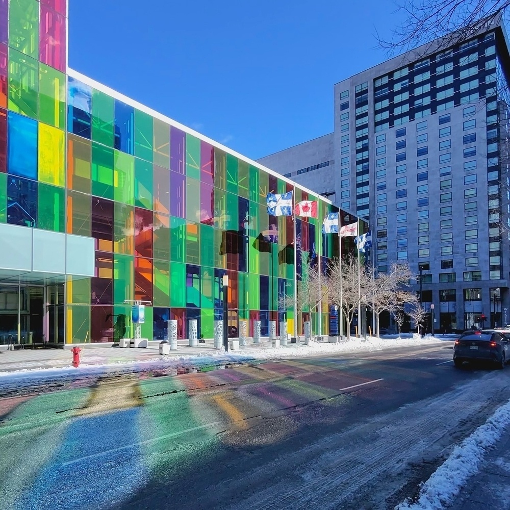 Colorful glass panels reflect vibrant patterns across a snowy urban street, with a modern building facade in the background. Flags flutter alongside bare trees under a clear blue sky.