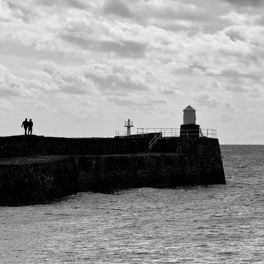 Black and white photo of a stone pier with two walking figures and two lighthouses, one mid-way and one at the end. 