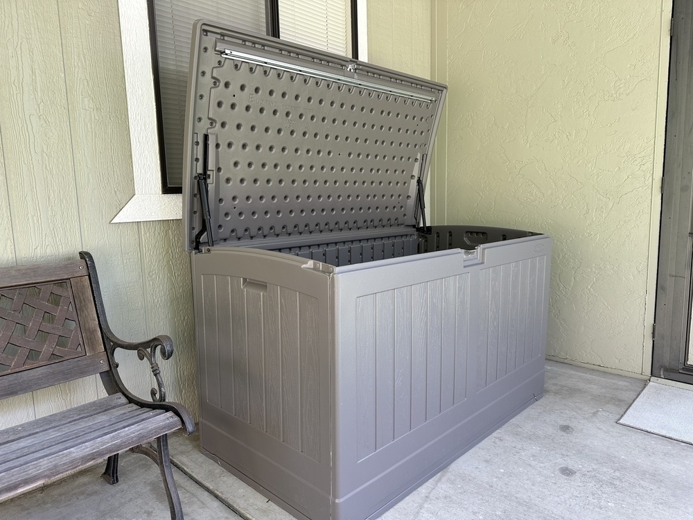 A large gray outdoor storage box with an open lid is placed next to a wooden bench on a patio.