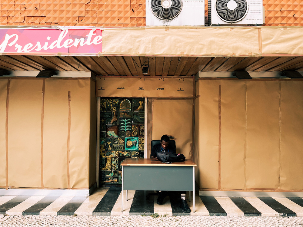 A man sits at a table, in front of a store that’s closed for renovation. 