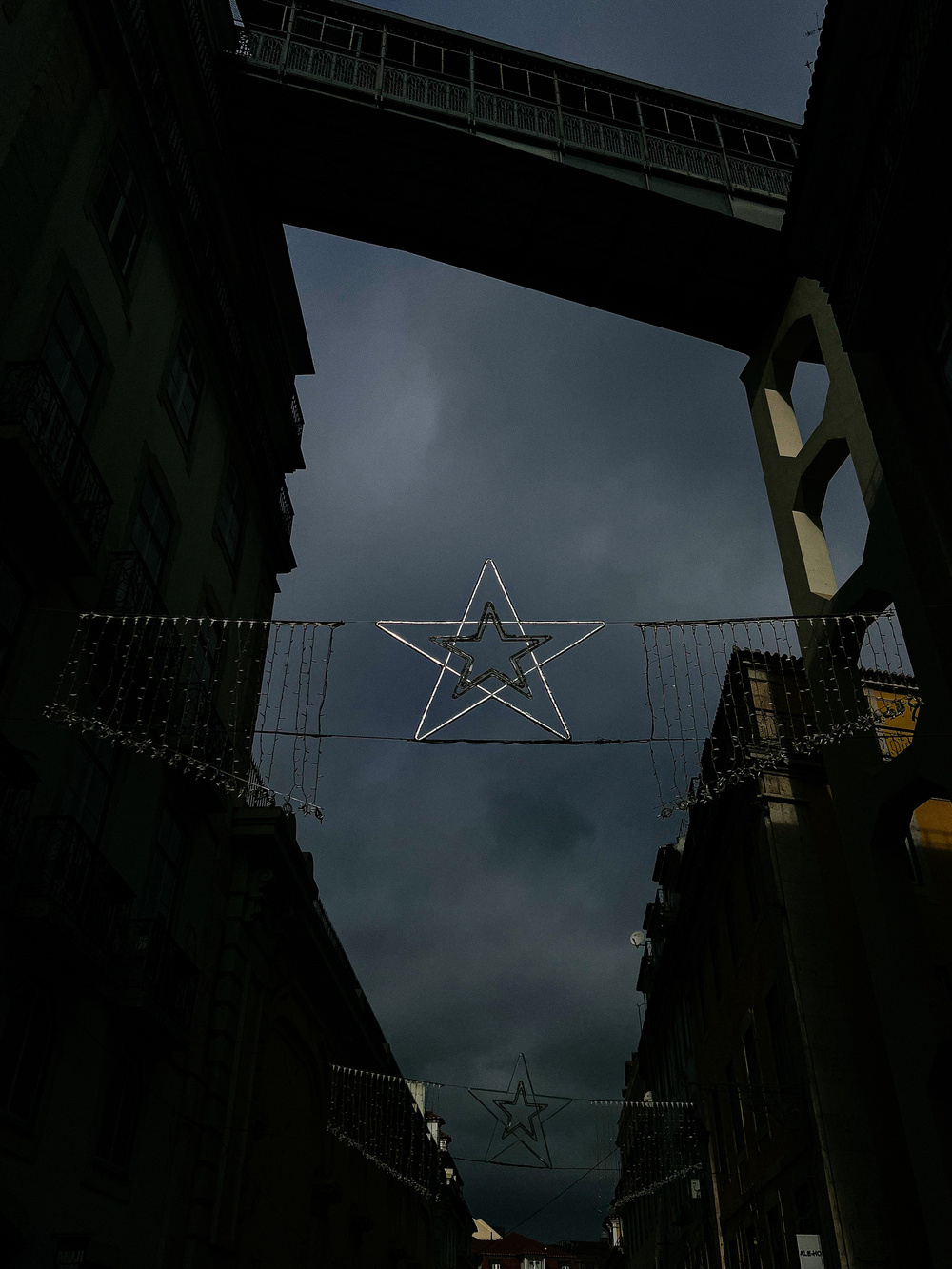 A narrow street flanked by multi-story buildings with balconies under an overcast sky. An illuminated outline of a star hangs between the buildings, and a pedestrian bridge spans the gap overhead.