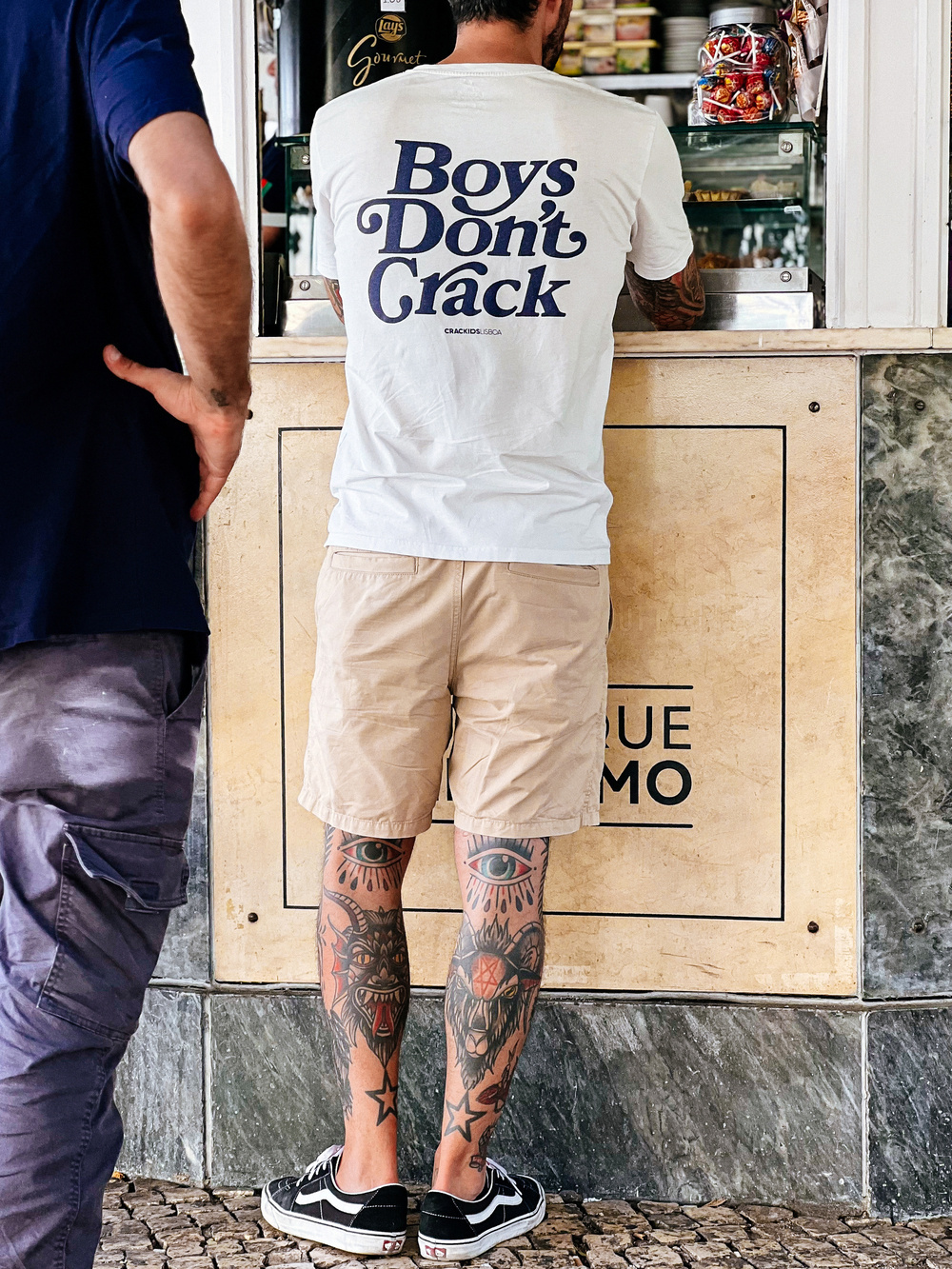 A tattooed gentlemen at a kiosk. His t-shirt reads “Boys Don’t Crack”. 