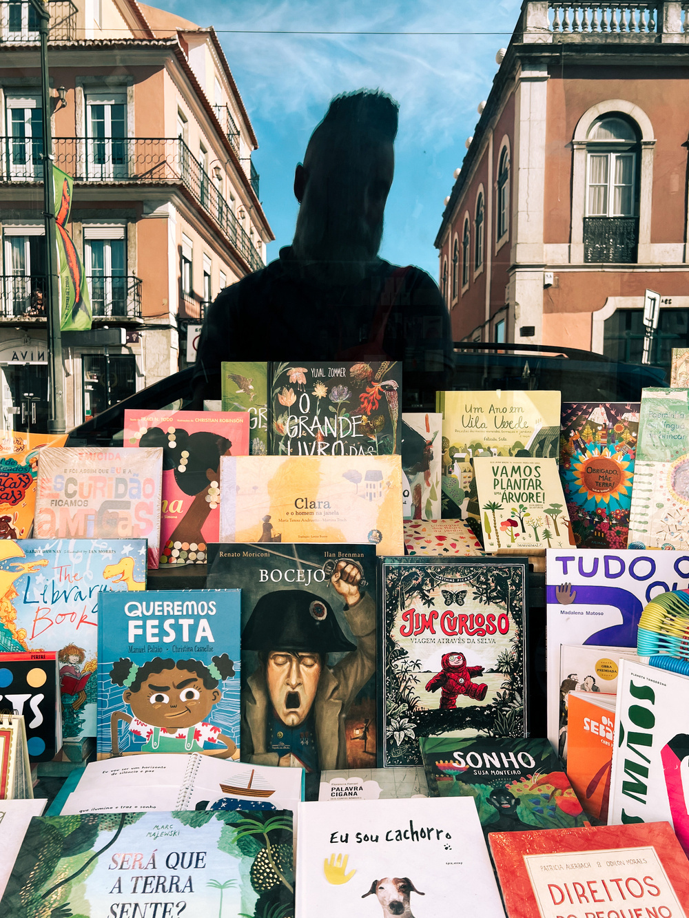 A man’s reflection on a bookstore window, filled with children’s books. 