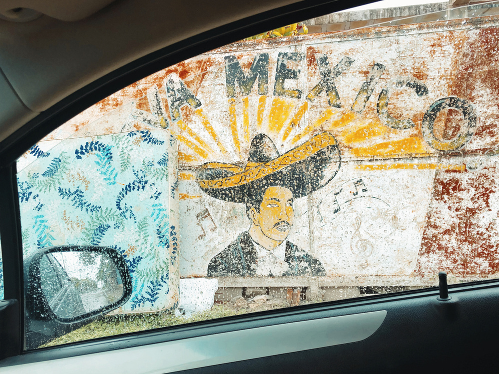 View through a car window on a rainy day, showing a mural outside featuring a man wearing a traditional Mexican sombrero with “VIVA MEXICO” written above him. There is also a blue, fern-patterned mattress beside the mural.