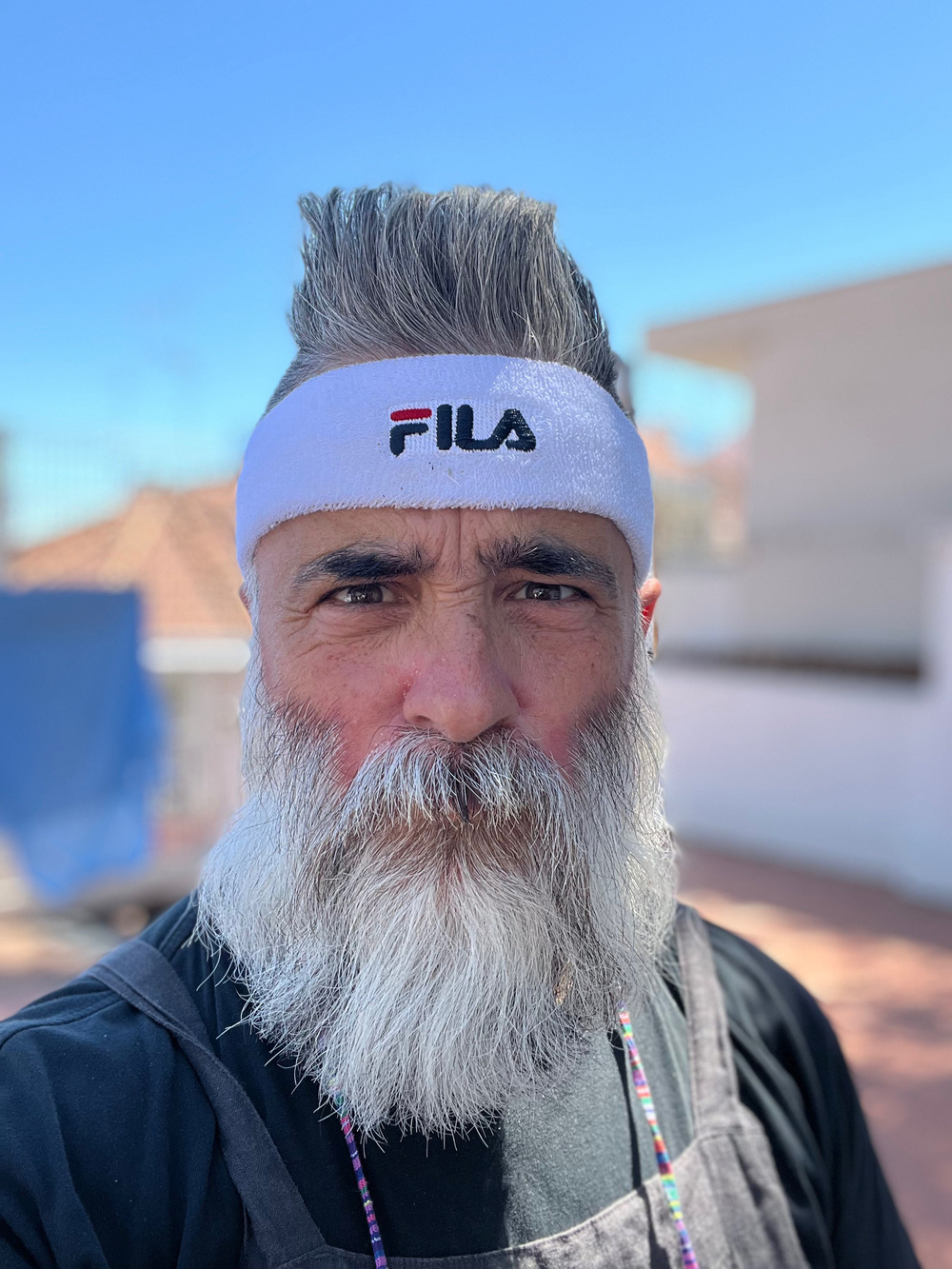 A man with a white headband featuring the &ldquo;Fila&rdquo; logo and a bushy white beard is standing outdoors. He is wearing a black shirt and appears to be in a sunny environment with a blue sky in the background.