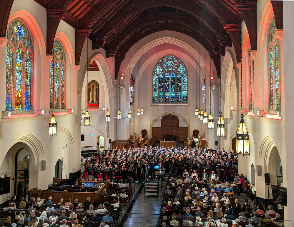 A large church with off-white walls and a dark-brown wooden ceiling. In the walls are complex and beautiful stained-glass windows. On the floor, the nearest third is occupied by the front rows of the audience. Then there is the very large choir, all in black, facing the camera; behind them the altar. The angle reveals some of the organ pipes facing us in the stage-right apse. Lights, framed in iron, hang on long cords from the ceiling, illuminating the choir and audience.