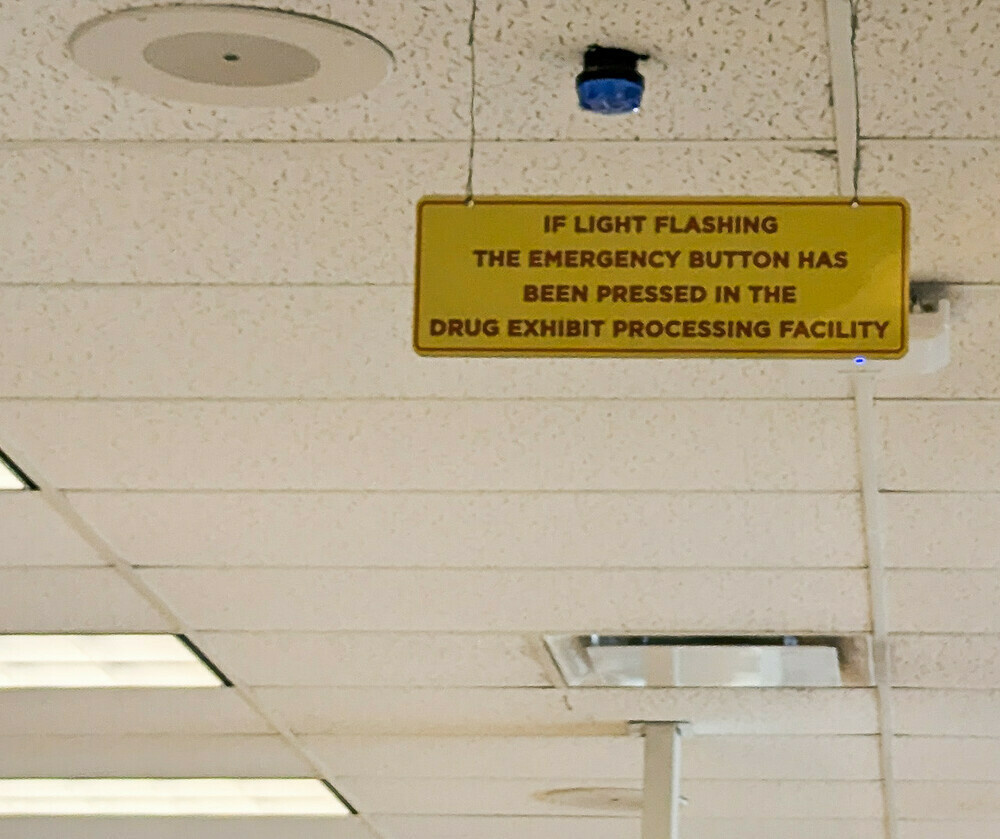 There's a blue light on an institutional ceiling; below it, a dark-red-on-yellow sign reads 
IF LIGHT FLASHING
THE EMERGENCY BUTTON HAS 
BEEN PRESSED IN THE
DRUG EXHIBIT PROCESSING FACILITY