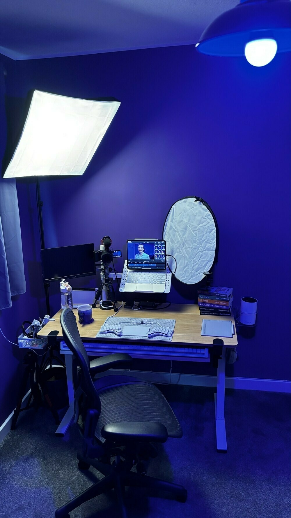 Desk with large soft box key light mounted to left, high up and angled downwards and inwards at forty-five degrees. Reflector on right, similar angle. Overhead light is a 40% intensity blue smart bulb.