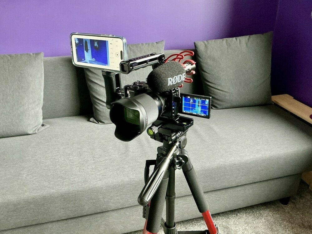 Mirrorless camera on tripod. The camera is in a cage, with a side handle mounted which has a MagSafe phone holder on top, holding my iPhone, which is set up as a remote monitor and control for the camera. The camera’s flip-out screen is deployed on the opposite side, and the cage has a top handle installed, on which a Rode shotgun mic is mounted.