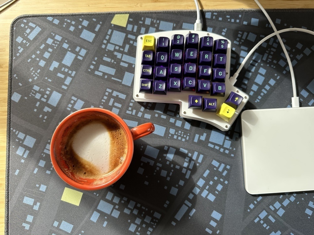 A top down view of the left half of a split keyboard, an Iris, with blue keycaps and acrylic white top case. Beside it sits an orange coffee cup. Both rest on a desk mat of dark blue with abstract lighter blue shapes.