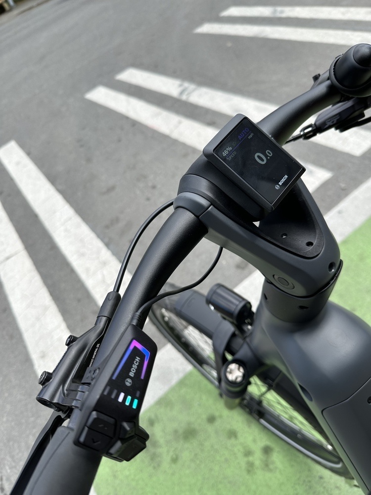 Shiny black handlebars of an e-bike with a small digital display at the top of the stem. In the background is the green bike lane marking of an intersection where the bike is safely stopped.