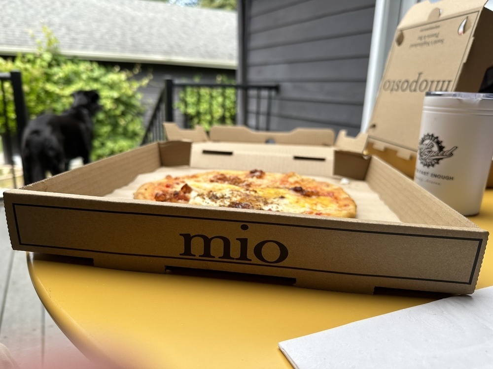 A pizza in a takeout box with the name ‘Mio’ printed on the edge of the box.