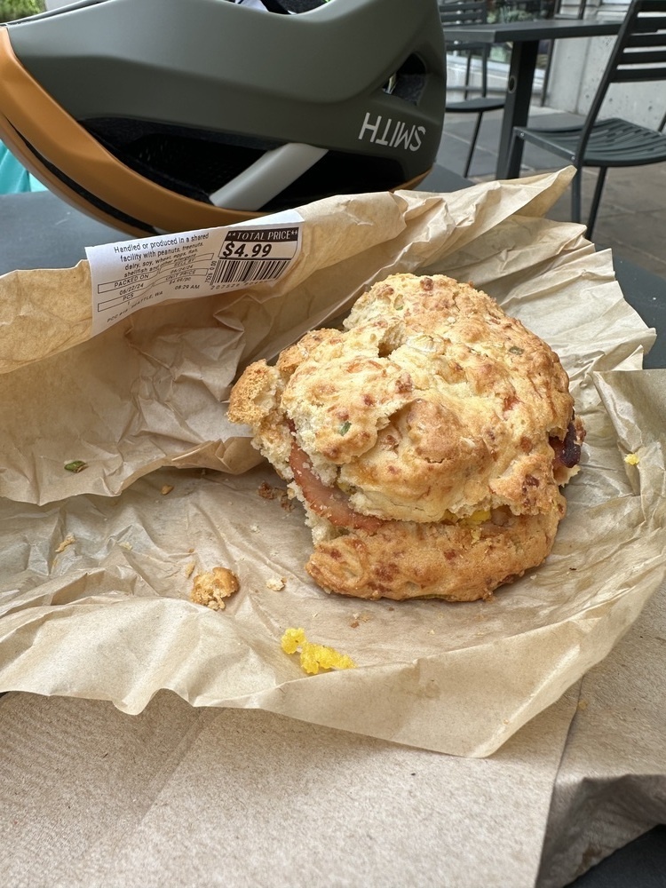 A breakfast sandwich on a big crunchy looking biscuit sits on a wrapper. Behind it is a gray and orange bike helmet. 