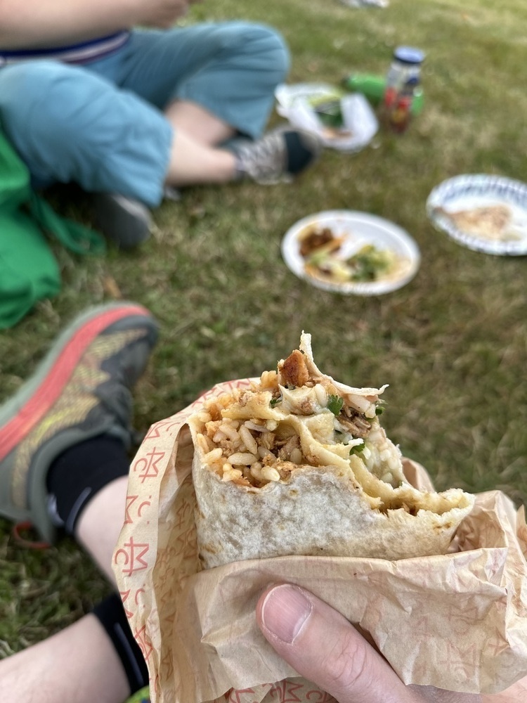 A half eaten burrito held in my hand, wrapped in brown paper. I’m sitting in the grass. 