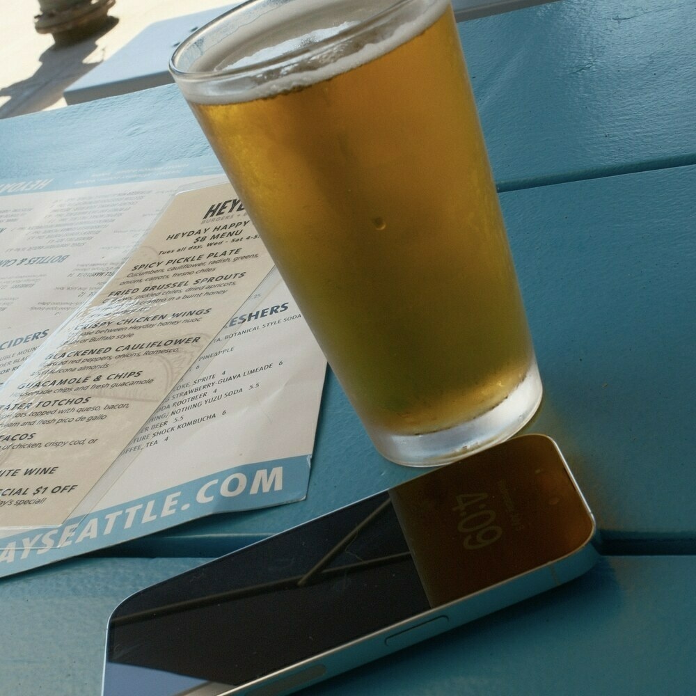 A golden-colored beer in a pint glass sits beside an iPhone 14 on a blue picnic table.