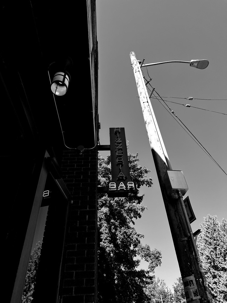 Monochrome photo of a telephone pole next to a sign reading pizzeria and bar. 