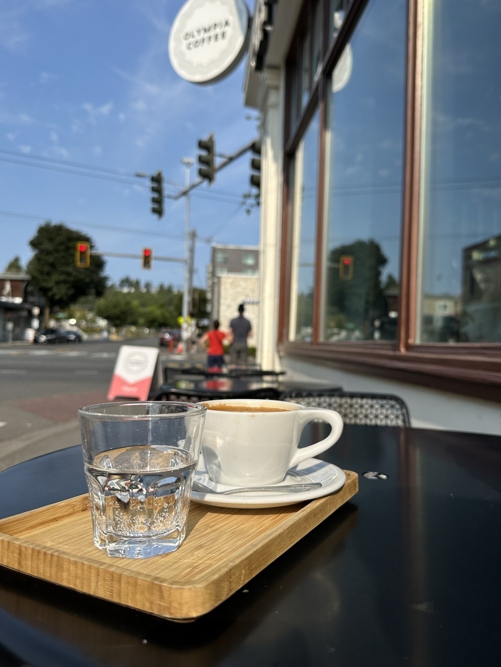 A cappuccino cup and a small glass of water sit on a bamboo tray, at a sunny sidewalk table.