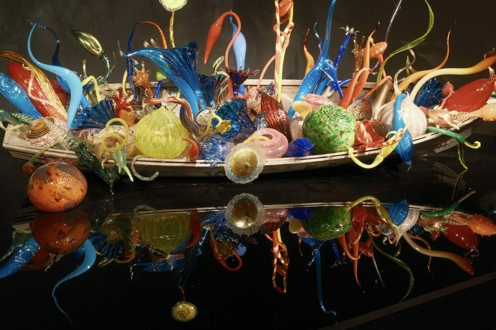 A wooden boat absolutely filled with multicolored glass balls and tendrils. The boat and its contents are reflected in a black glass platform beneath them.