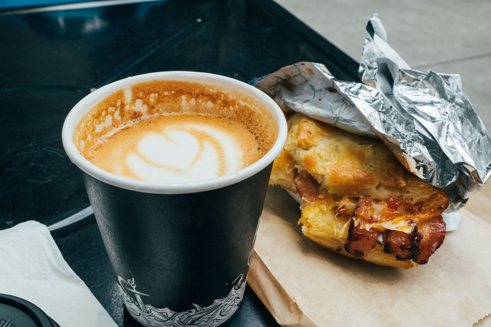 A cappuccino in a black paper cup. It is a rich brown color with a rose-shaped pour of milk on top. Next to it is a small biscuit sandwich with cheddar cheese, bacon, and pepper jam. Both of them look absolutely delicious.