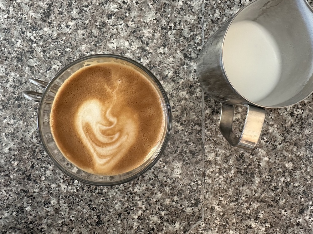 Top down photo of a cappuccino in a glass cup. It looks pretty good, smooth and a wavy mix of brown and white, despite the milk pour being messy. It sits on a speckled countertop beside a silver pitcher.