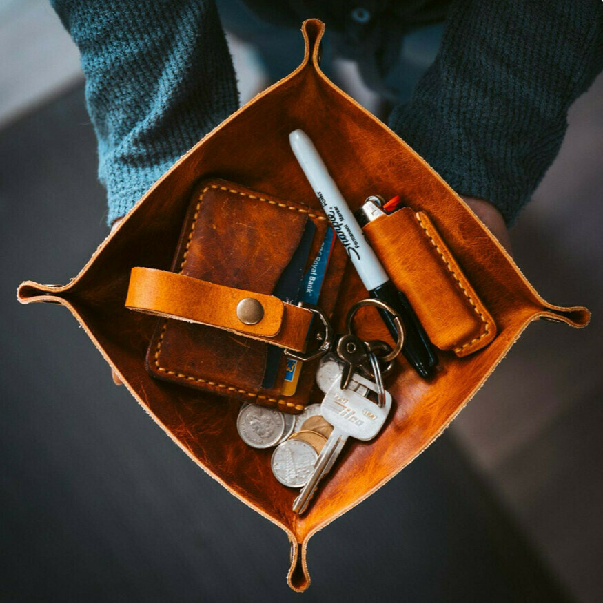 A picture from above of a person holding a leather valet tray in their hands. In the tray are several items: coins, keys on a leather keychain, a leather wallet, a black Sharpie marker, and a Bic lighter encased in a leather sheath.
