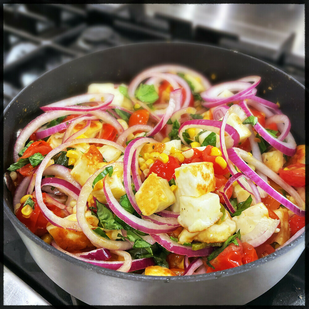 skillet filled with golden-brown chunks of halloumi, thinly sliced onions, halved cherry tomatoes, corn kernels, and chopped basil