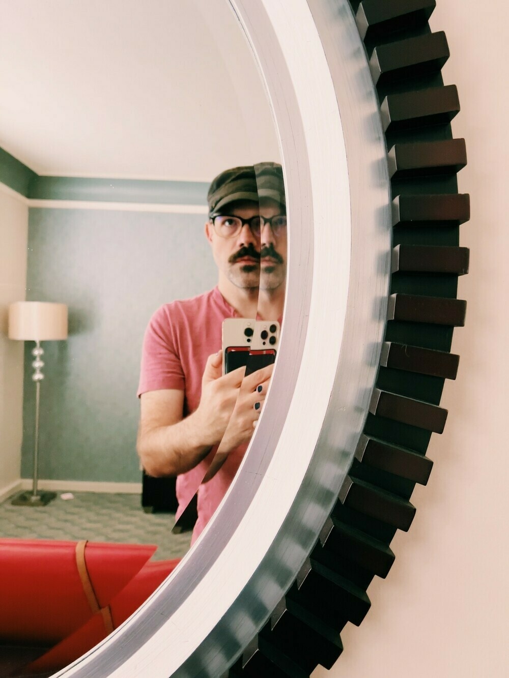 Self portrait. I’m wearing a black army cap, pink shirt, and glasses. I have a mustache and a little patch of fuzzy beard under my bottom lip. I’m holding an iPhone in front of a mirror and staring beyond the lens of the camera. My face is bisected by the curved mirror, refracting in a way that makes it look like it is splitting in two. 