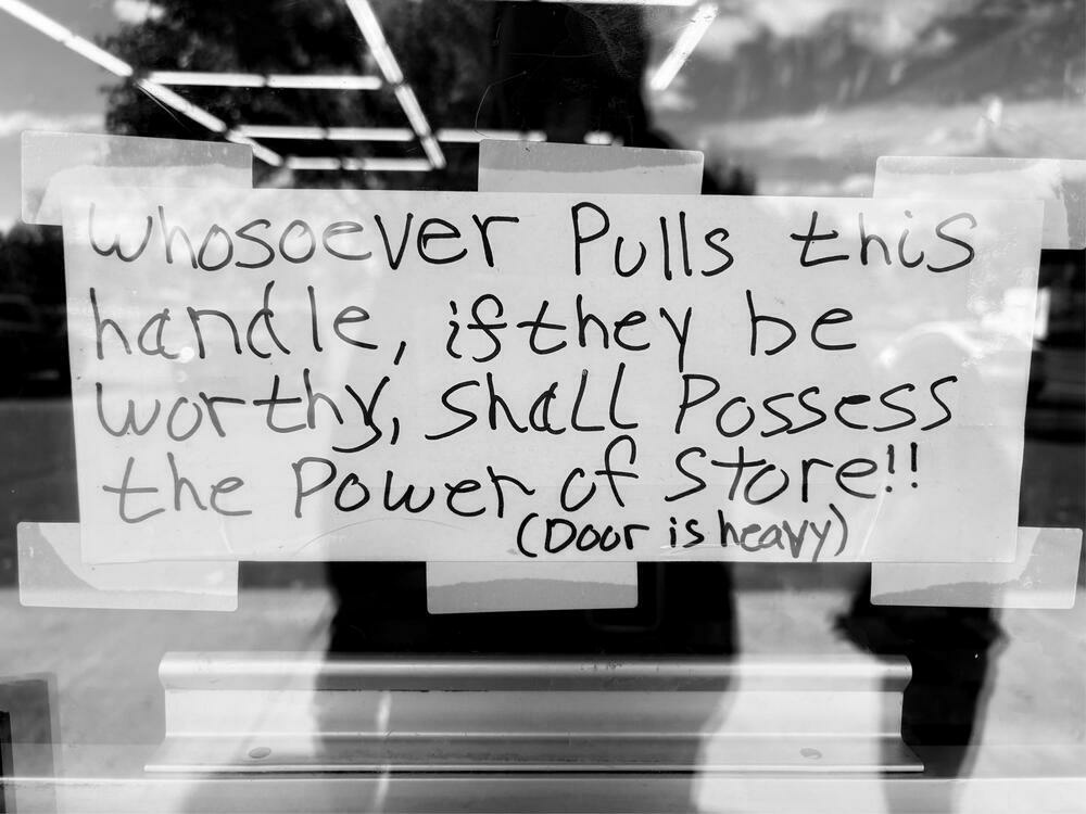 A humorous handwritten sign taped to a glass door reads, “Whosoever pulls this handle, if they be worthy, shall possess the power of store!! (Door is heavy).” The reflection of the parking lot and sky is visible in the glass behind it.
