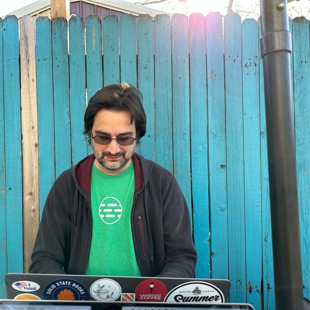 Manton at his computer at an outdoor coffee place, turquoise fence in background 