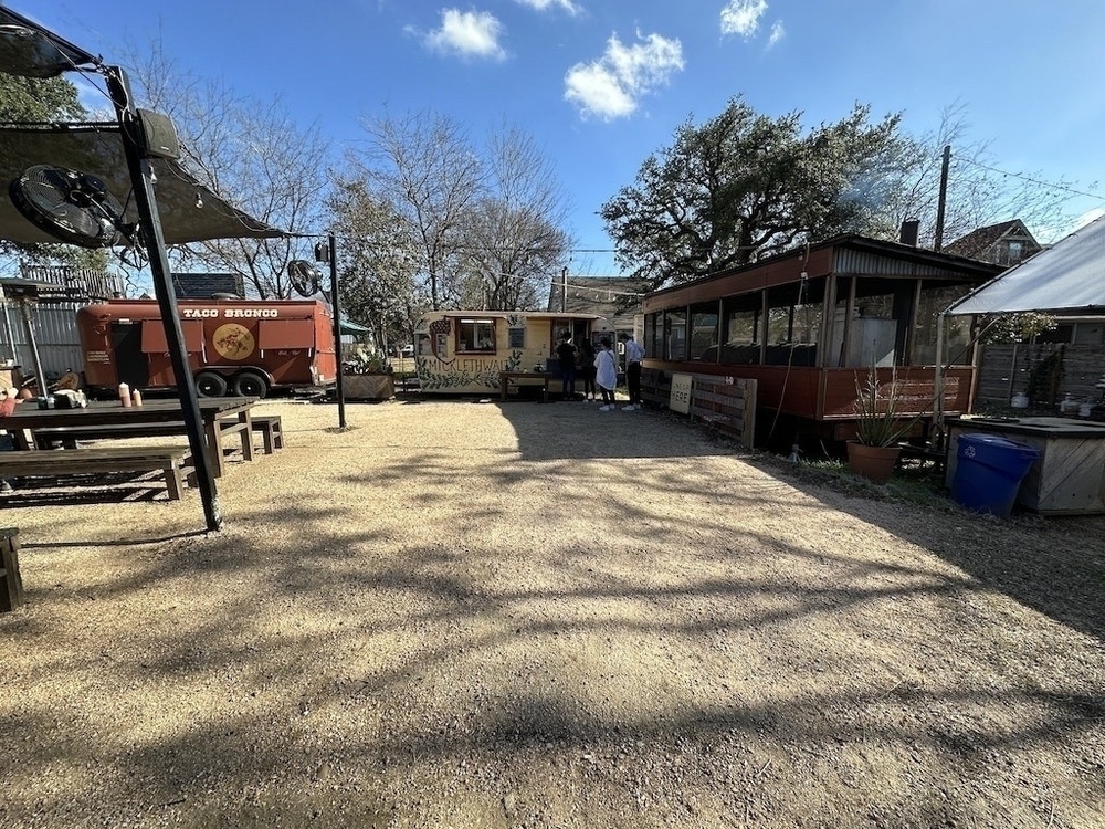 Wide angle shot with BBQ cart, Taco truck, picnic tables, gravel grounds, oaks in the background, blue sky with a few wispy clouds. 